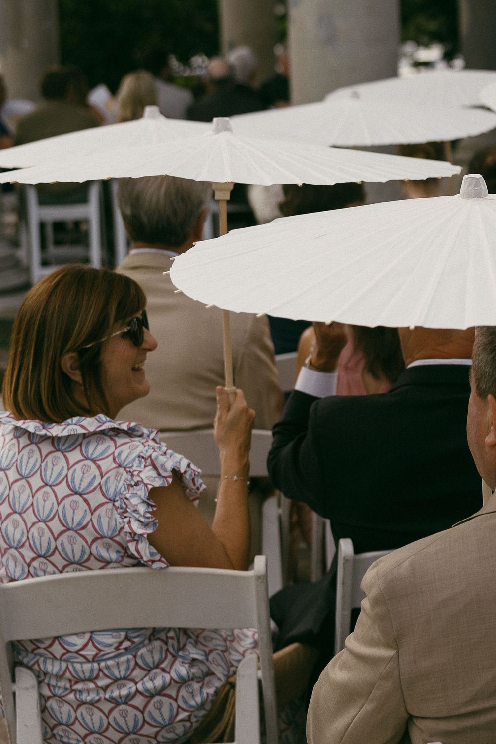 Guests seated outdoors holding white paper parasols at an elegant garden event on a sunny day.