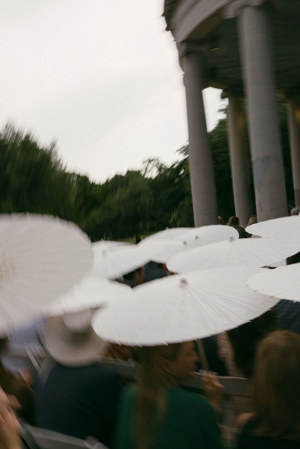 Blurred crowd holding white parasol umbrellas near a classical columned building with trees in the background.