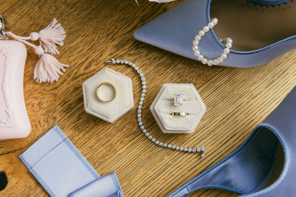 Elegant bridal flat lay featuring diamond rings in velvet boxes, pearl bracelet, blue heels, and pink tassel clutch on wood.
