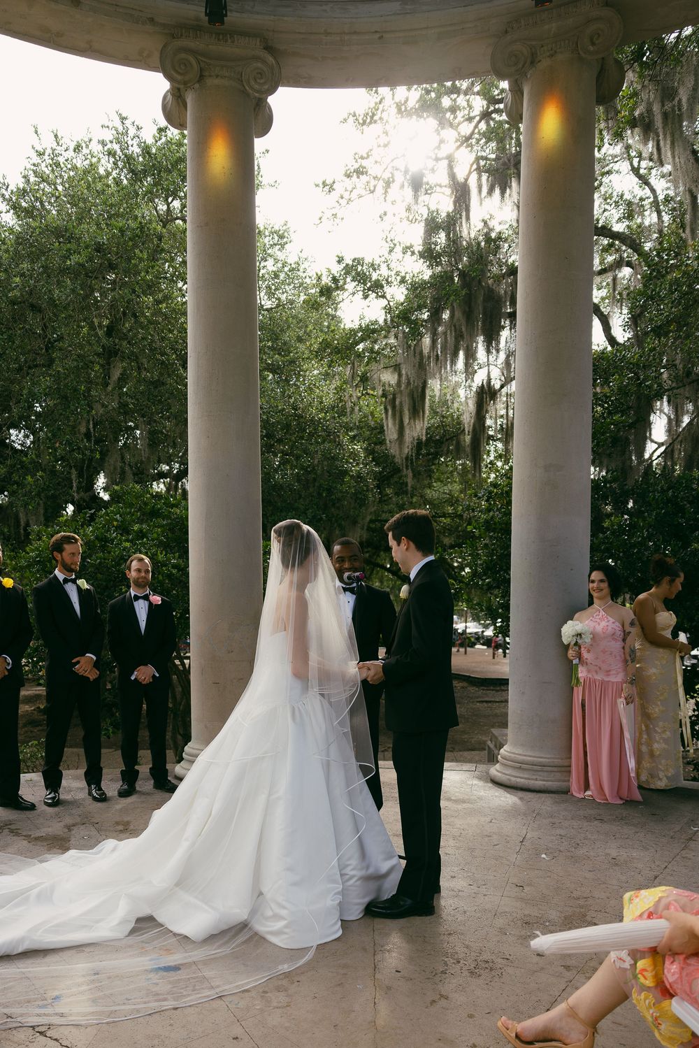 Bride and groom exchange vows under elegant stone columns at an outdoor wedding ceremony with lush greenery.