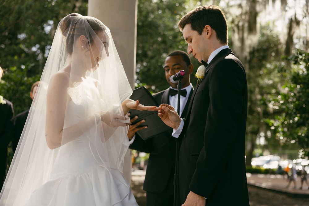 Bride and groom exchanging rings during outdoor wedding ceremony with officiant in black tux amid lush greenery.