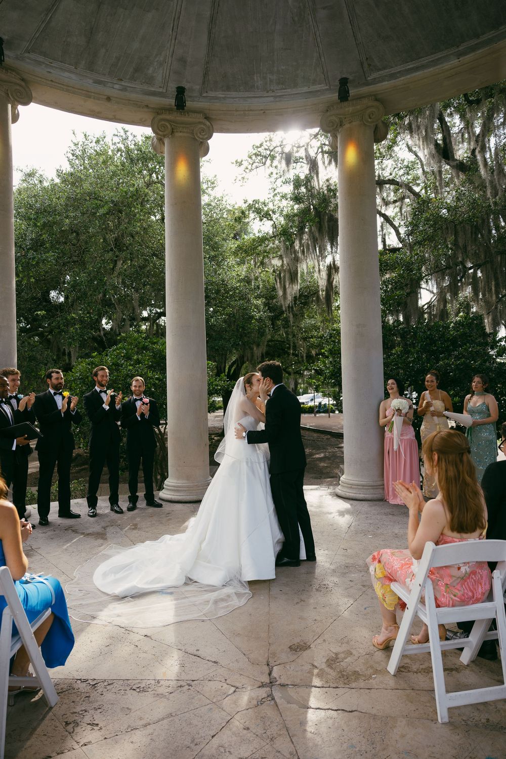 Bride and groom share first kiss under elegant stone gazebo with columns at outdoor wedding ceremony.
