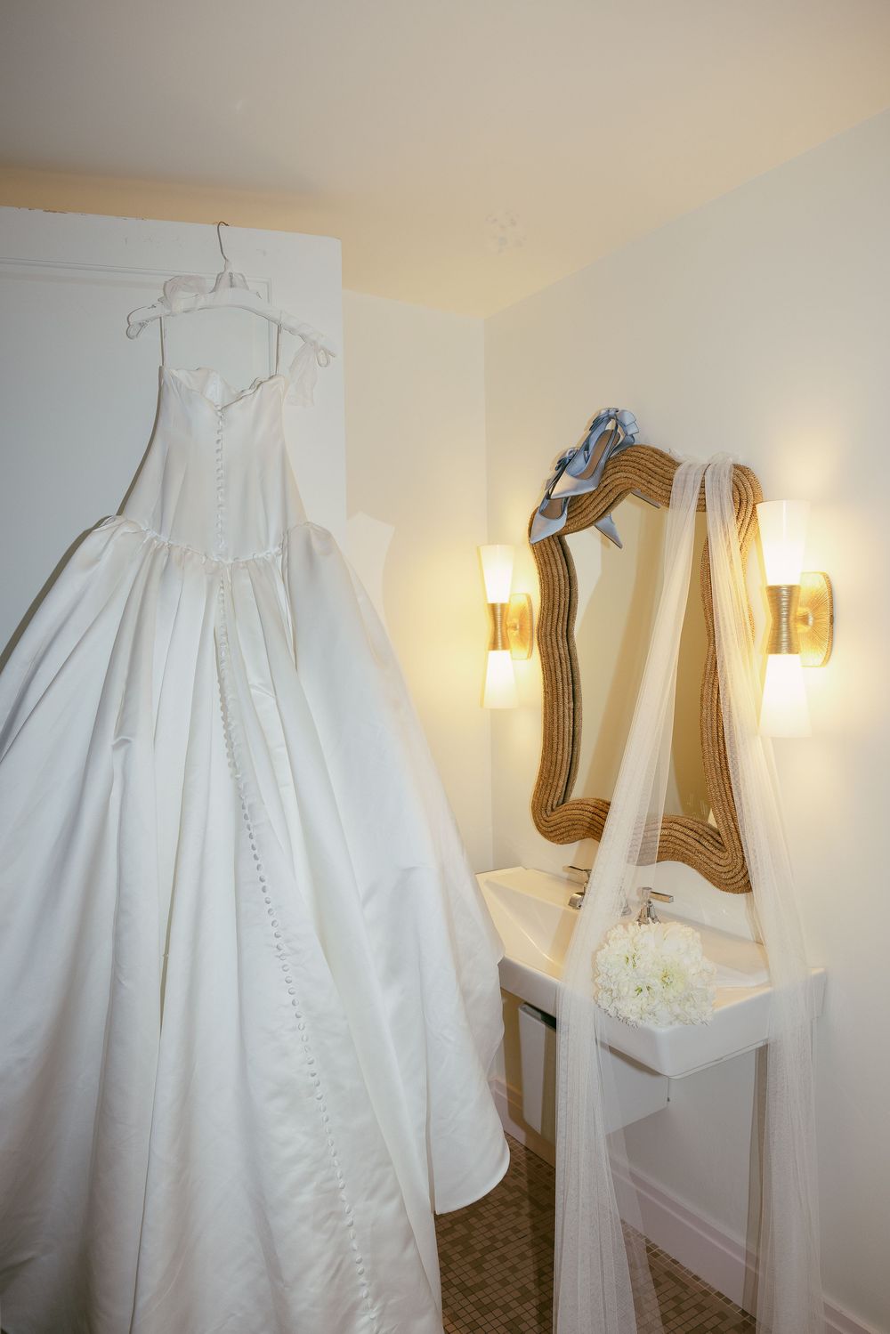 Elegant white ball gown wedding dress hanging in a bridal suite beside a gold mirror with veil and floral bouquet.
