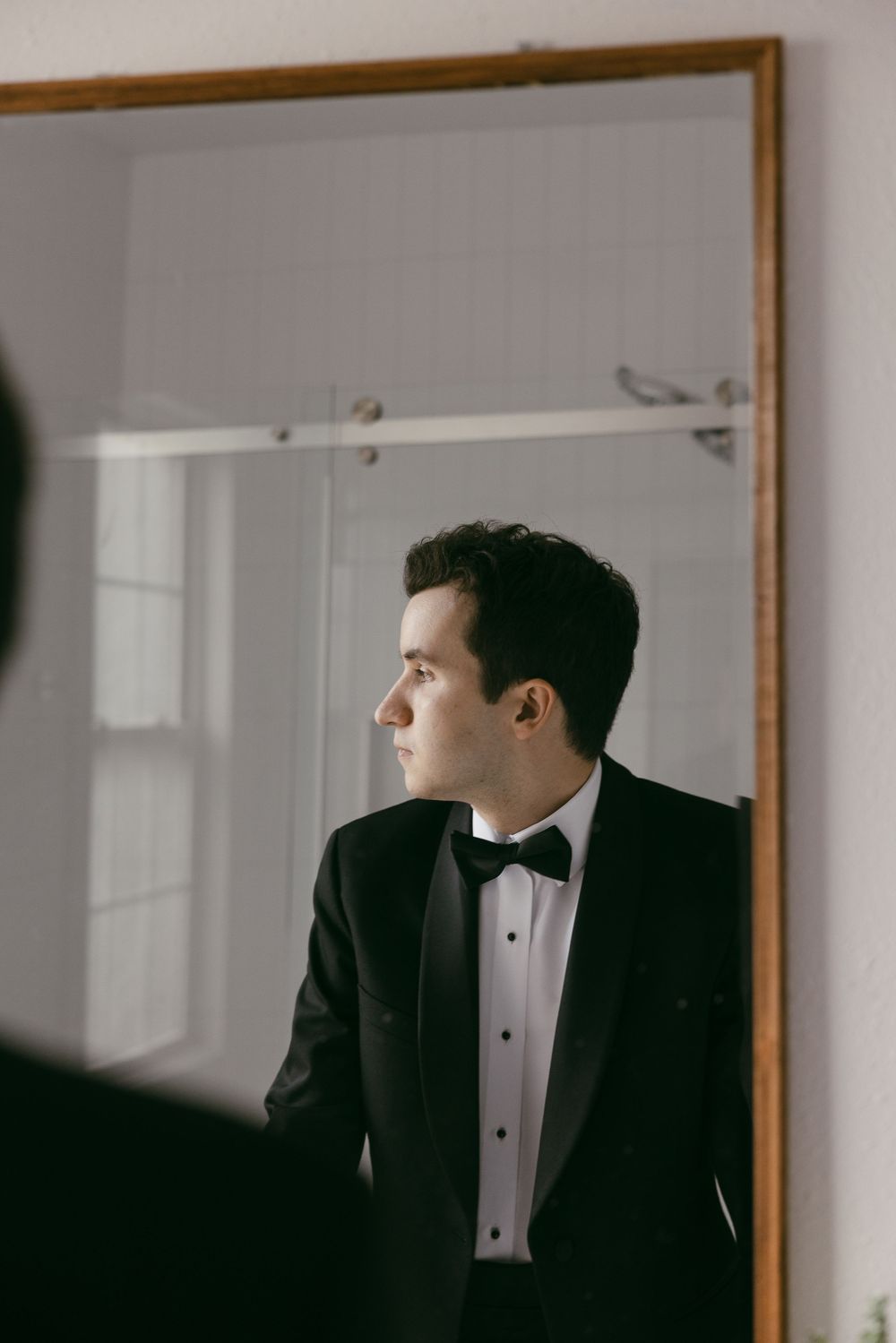 A groom in a black tuxedo and bow tie gazes sideways near a large mirror in a softly lit room.