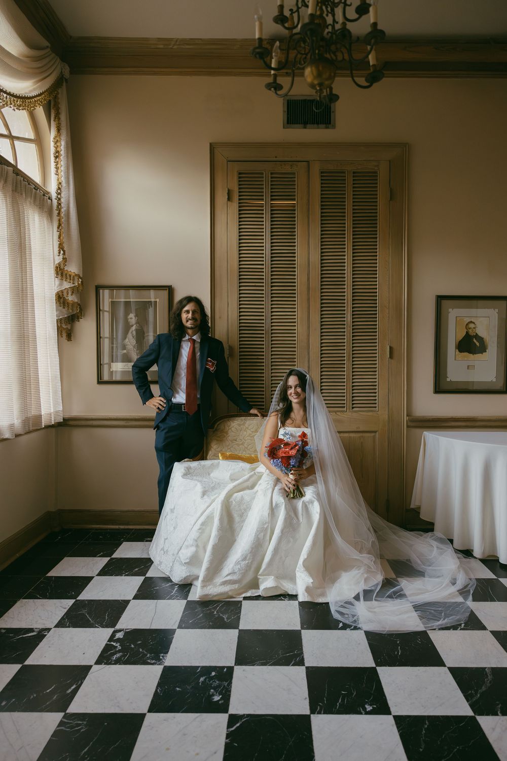 Bride in white gown with veil and bouquet sits while groom in navy suit stands on black and white checkered floor.