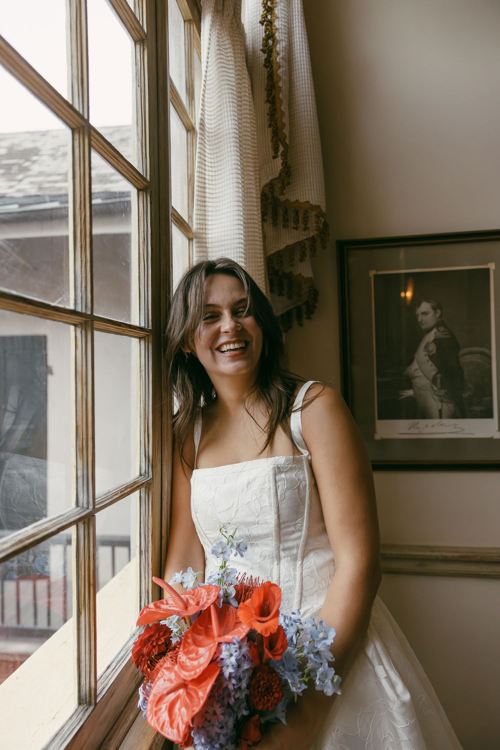 Smiling bride in white dress holding colorful bouquet of red and blue flowers, standing by a window in elegant indoor setting.