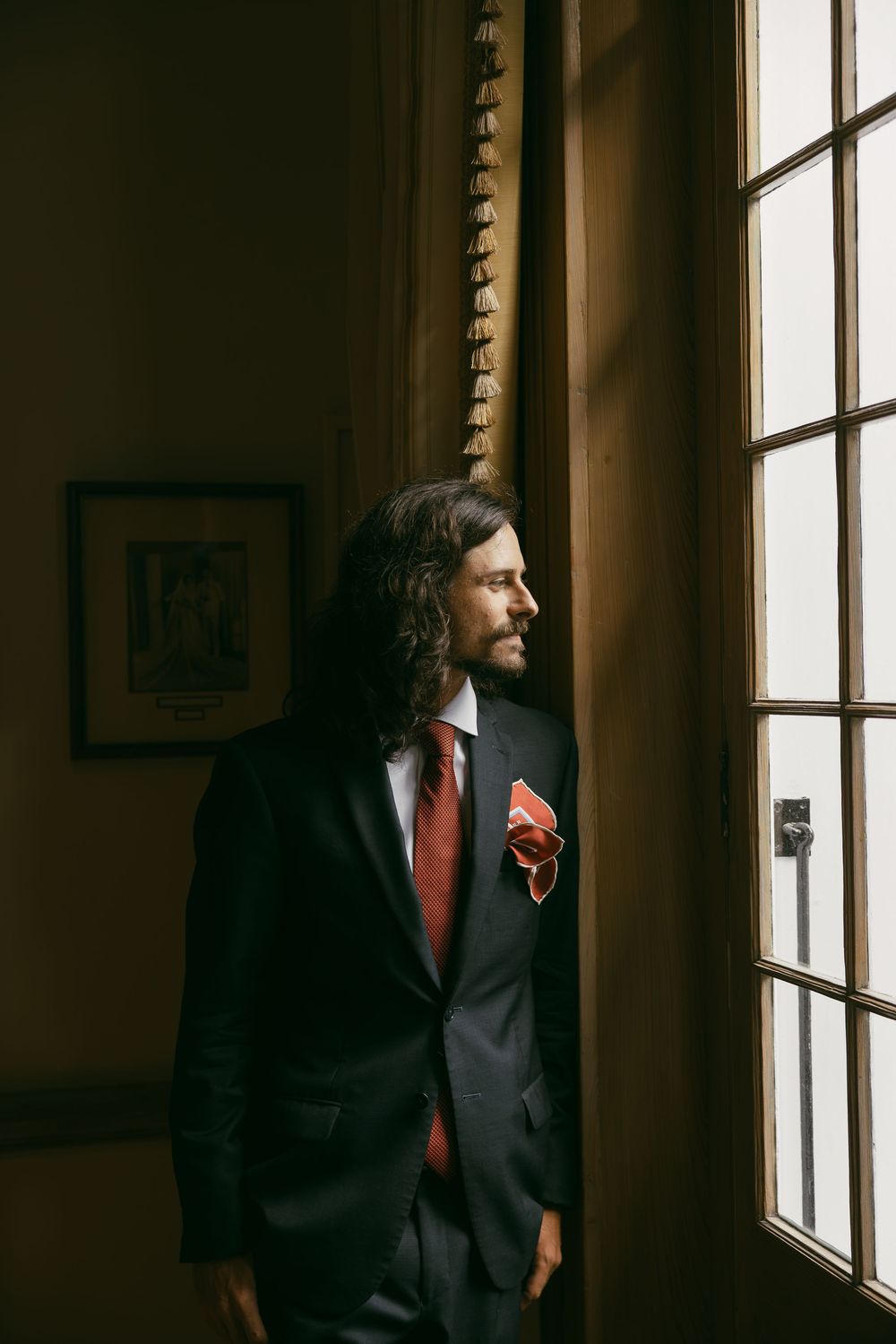 A bearded man with long curly hair in a dark suit, red tie, and pocket square stands by a sunlit window.