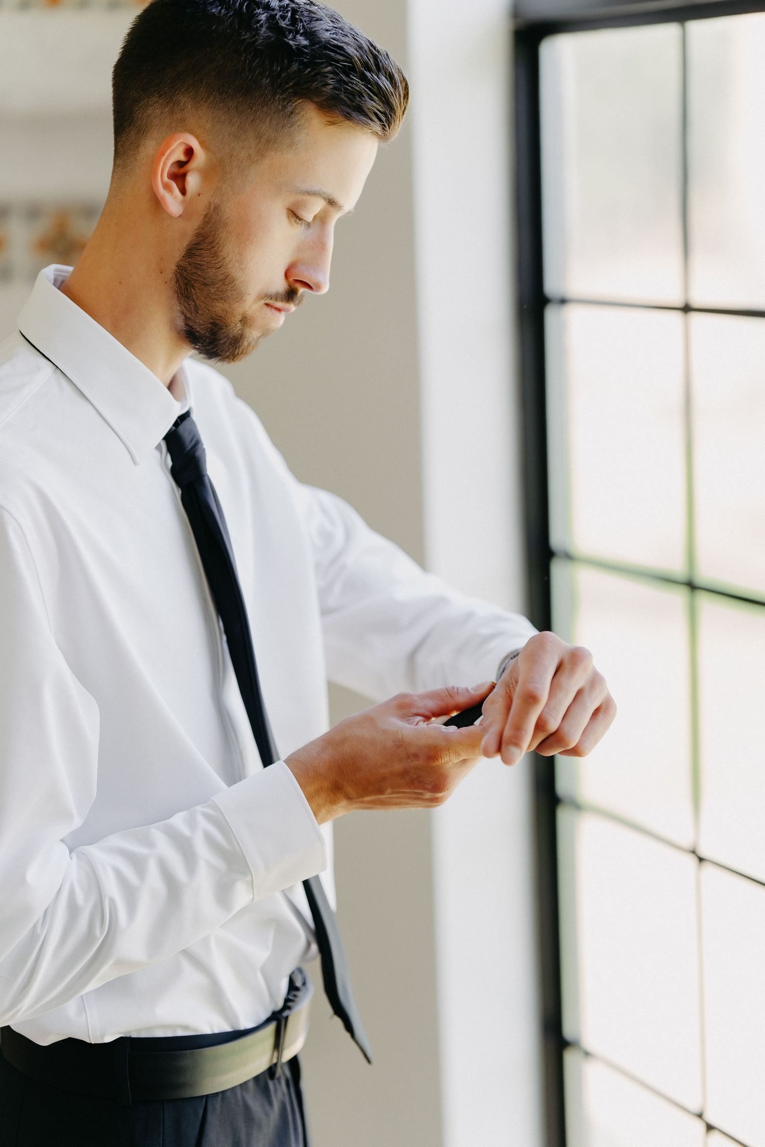 Groom adjusts cufflinks near bright window in formal attire.