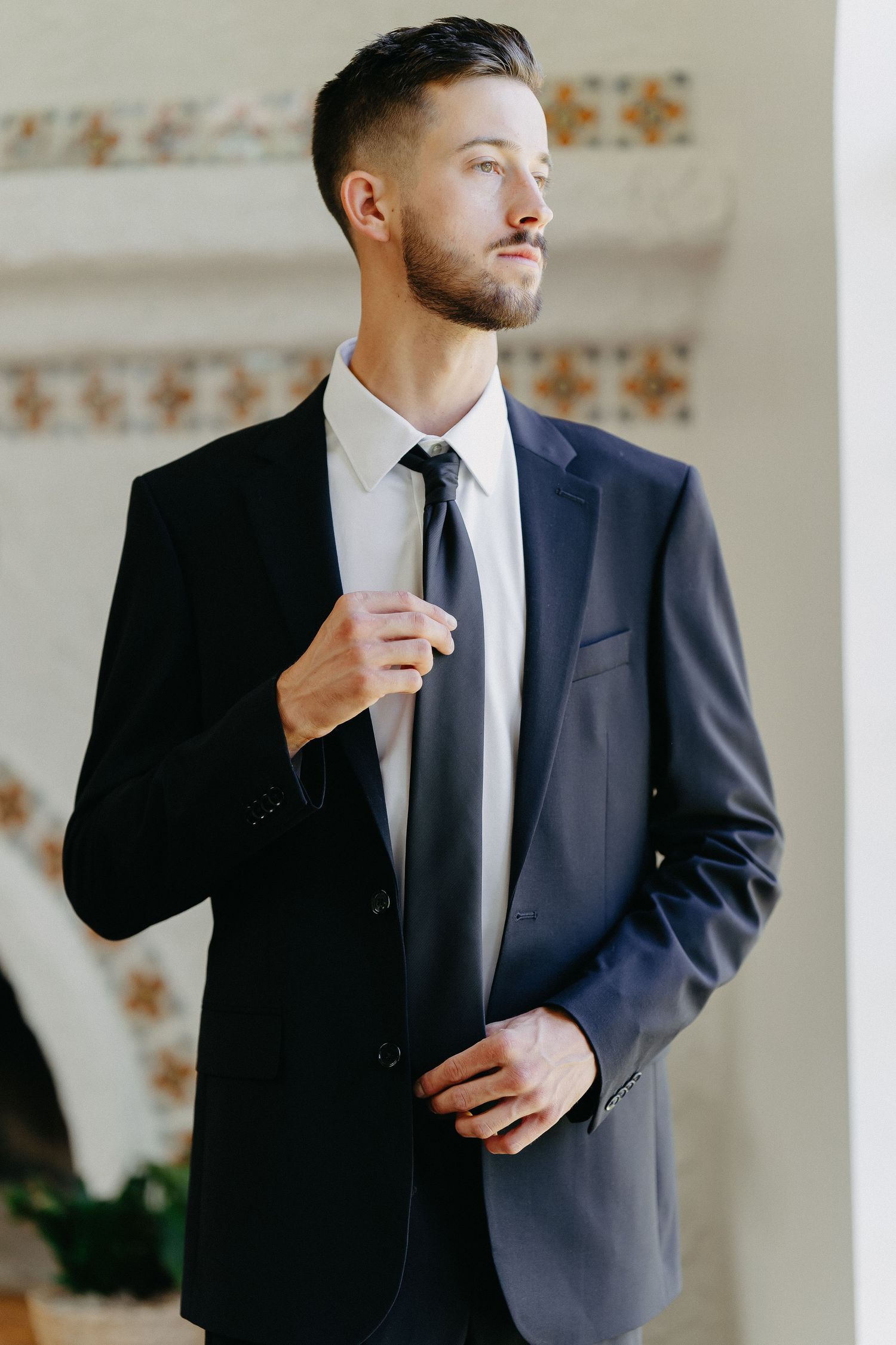 Classic portrait of groom in navy suit adjusting tie by window.