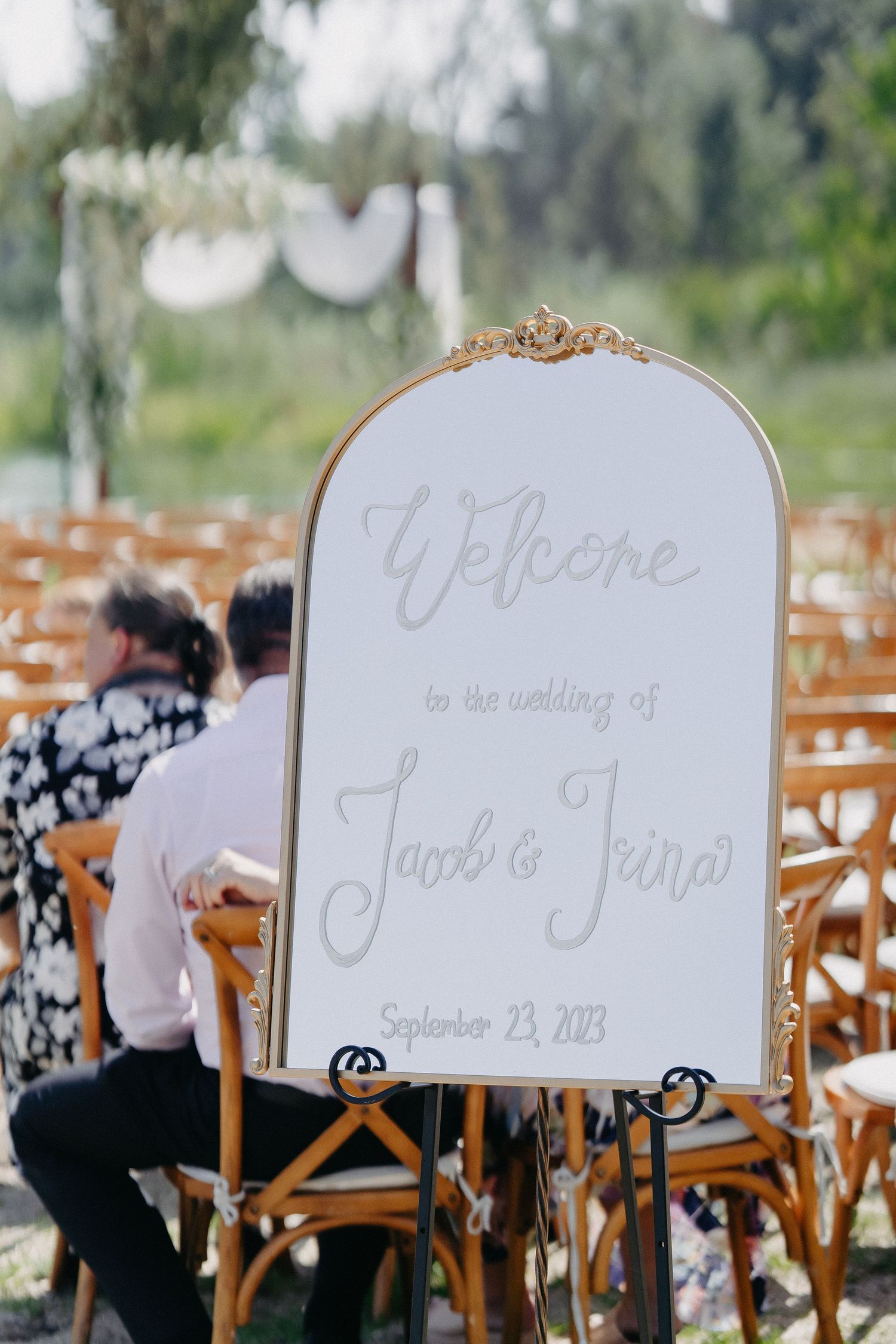 Elegant mirror welcome sign displayed on wooden easel at outdoor wedding venue.