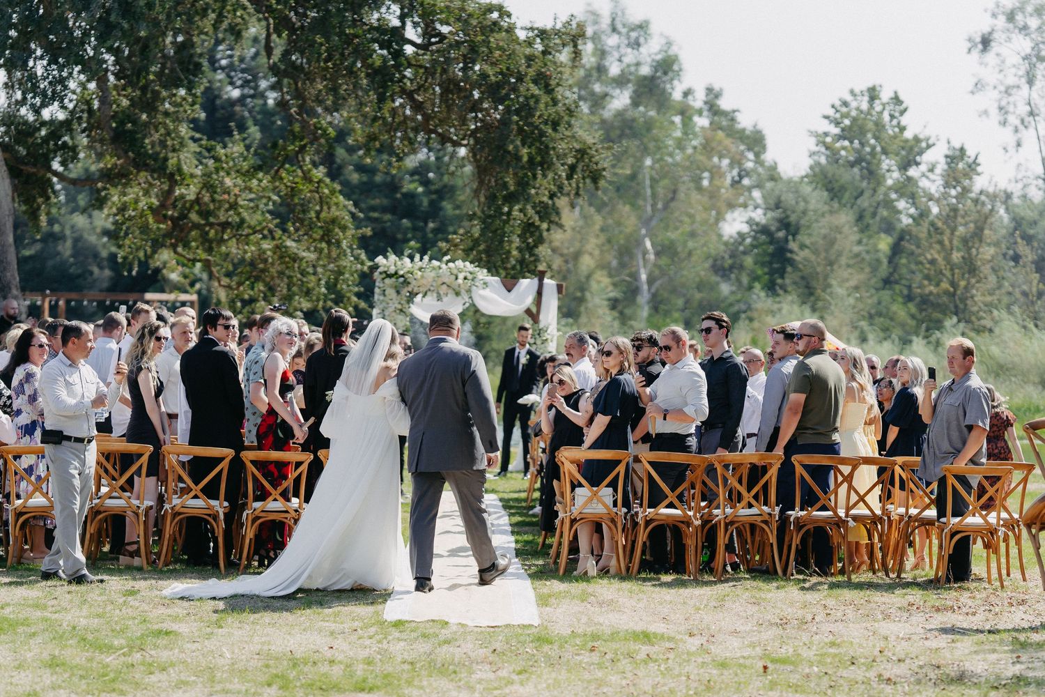 A wedding ceremony takes place outdoors with wooden crossback chairs arranged in rows on grass under trees.