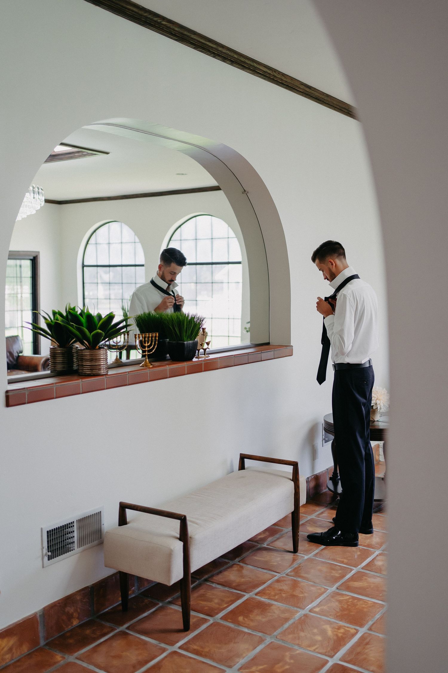 Two men in formal wear preparing in Spanish-style interior with arched windows.