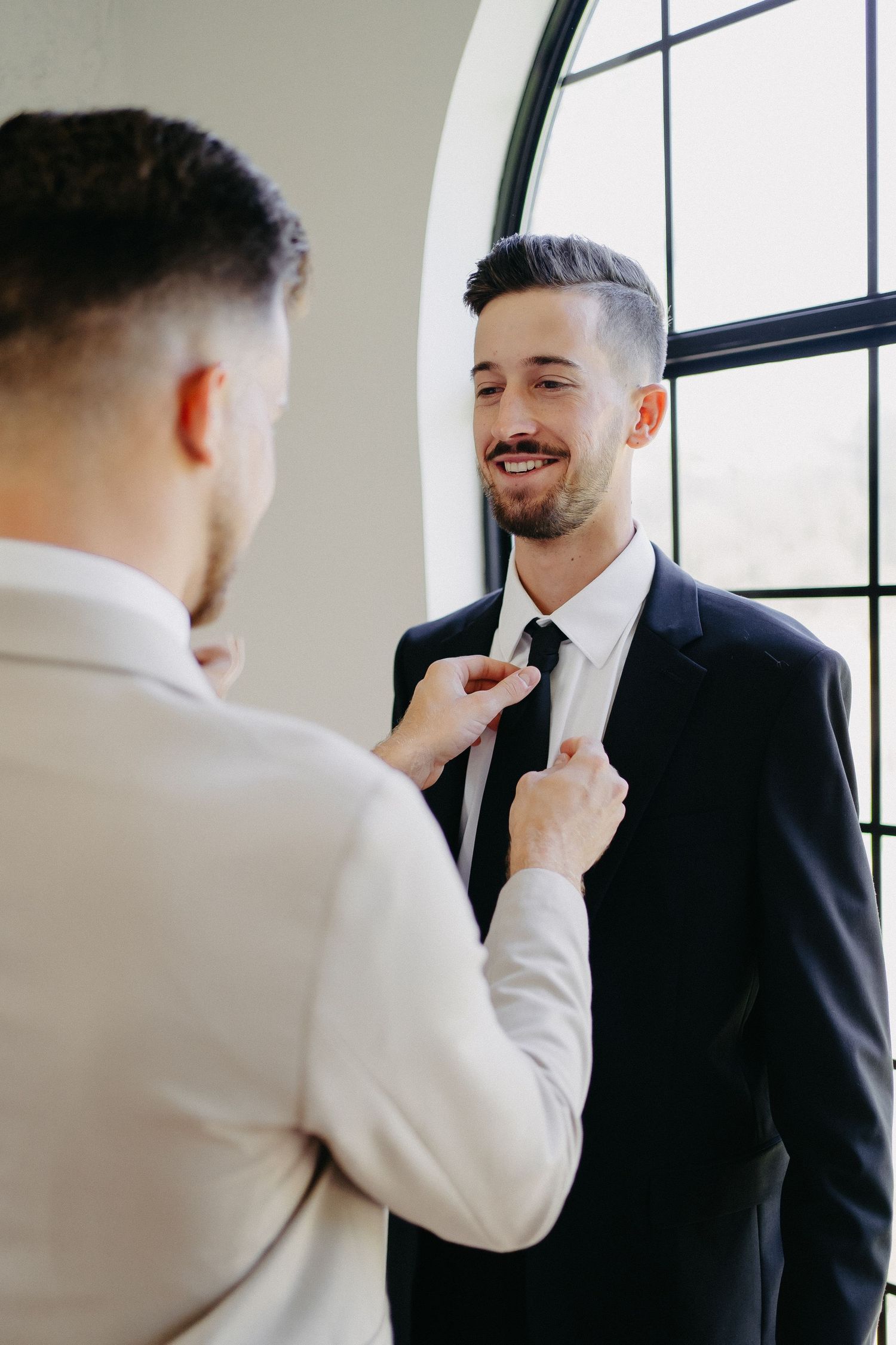 Two people in suits adjusting a necktie during wedding preparations near a large window.
