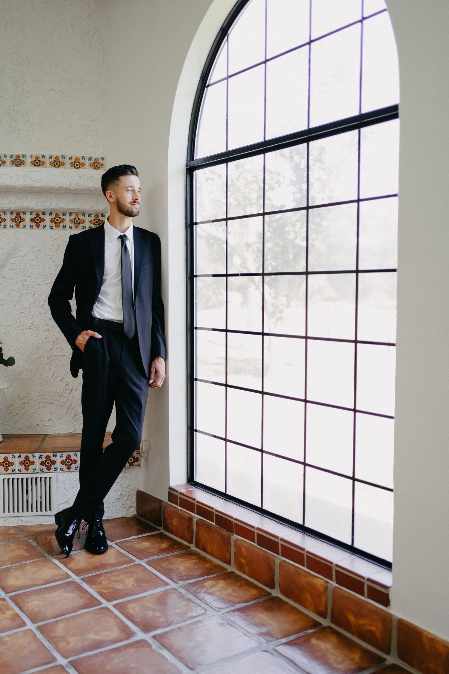 Well-dressed person standing near an elegant arched window in a Spanish-style interior.