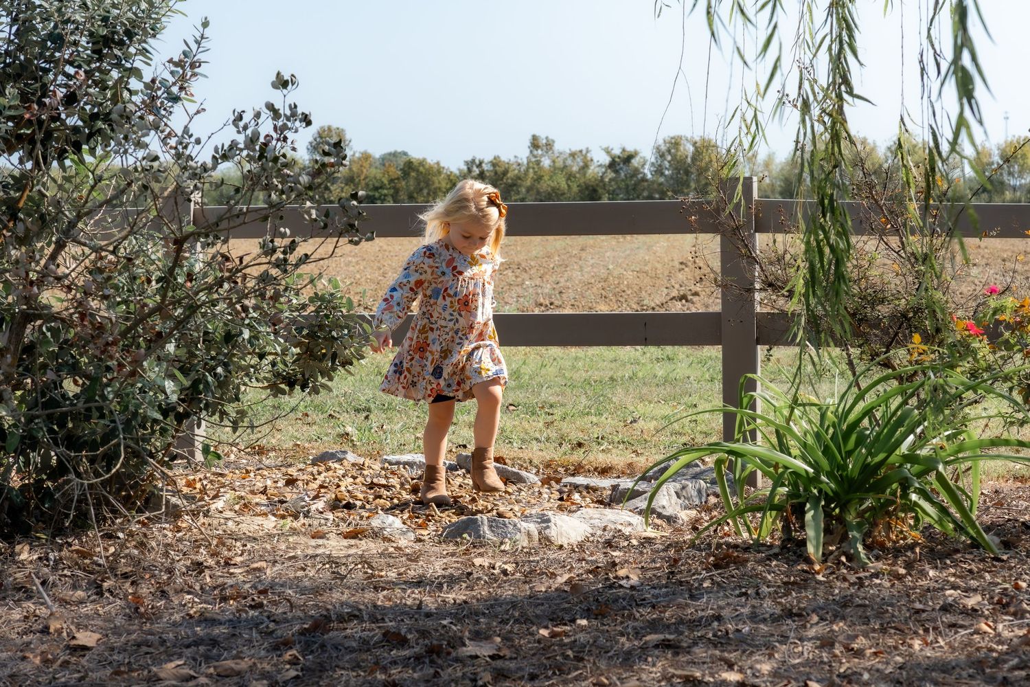 Young child exploring garden path under willow trees in a rustic country setting.