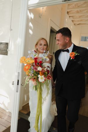 Newlywed couple sharing a romantic moment in a bright hallway with the bride holding a vibrant orange and red bouquet.
