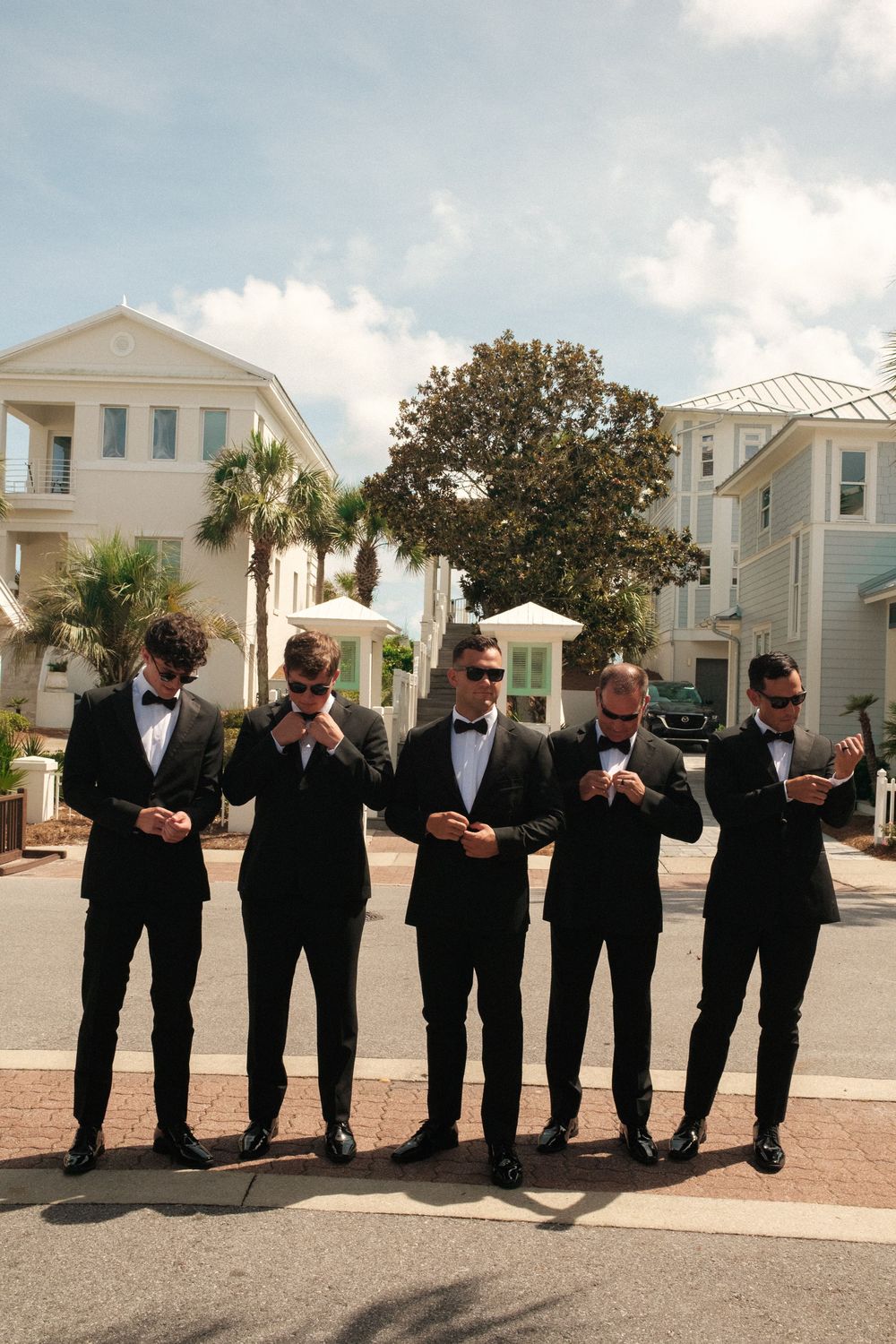 A line of groomsmen in black tuxedos check their watches in synchronized poses outside.