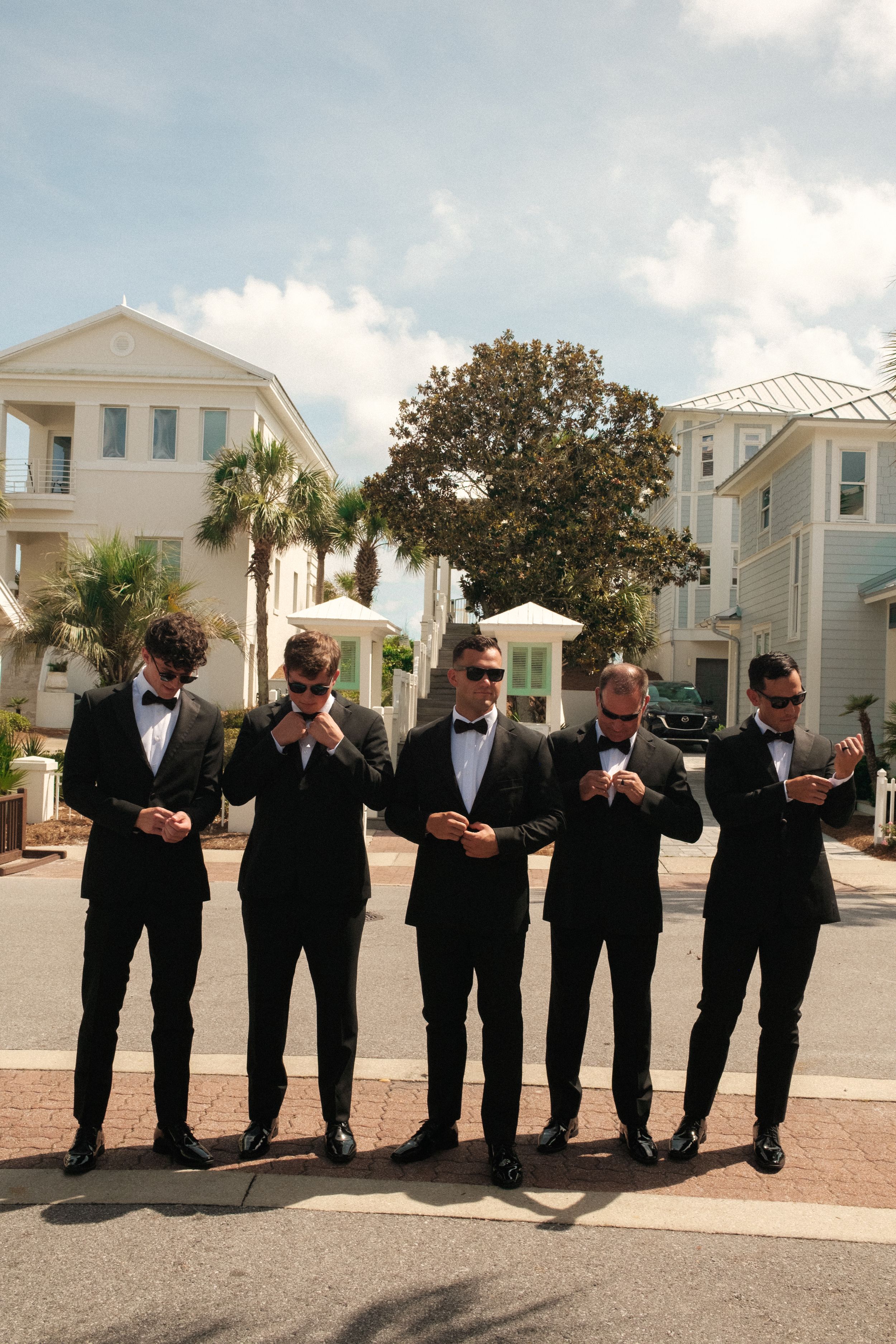 A line of groomsmen in black tuxedos check their watches in synchronized poses outside.