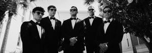 Groomsmen in black tuxedos posing together outdoors in black and white photography.