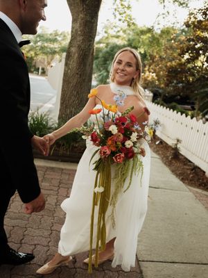 A couple in formal wedding attire poses outdoors with a vibrant red and white floral bouquet.