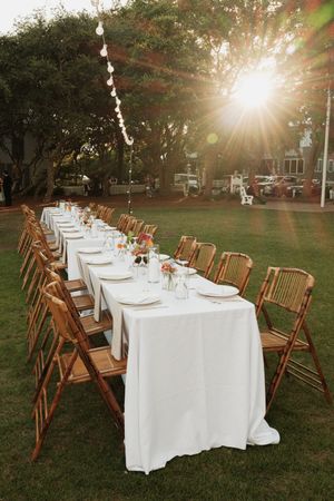 Outdoor wedding reception tables set with white linens and wooden chairs under trees at sunset.