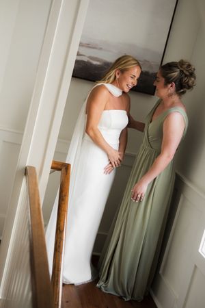 Two people in white and sage green dresses share a candid moment in a bright room.