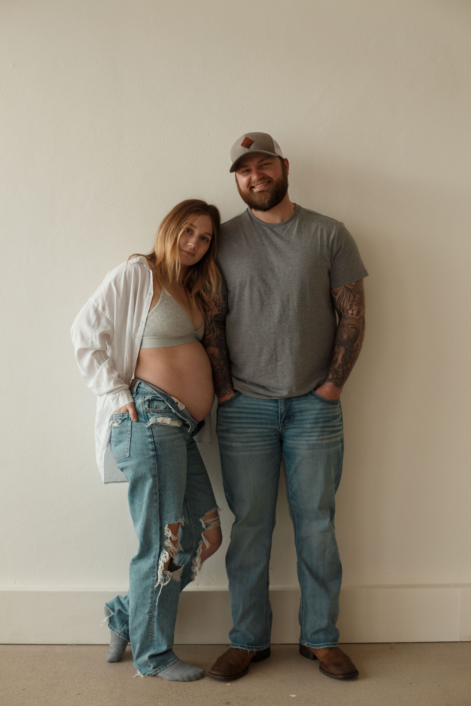A couple stands together against a white wall, wearing casual denim and displaying a visible pregnancy bump.