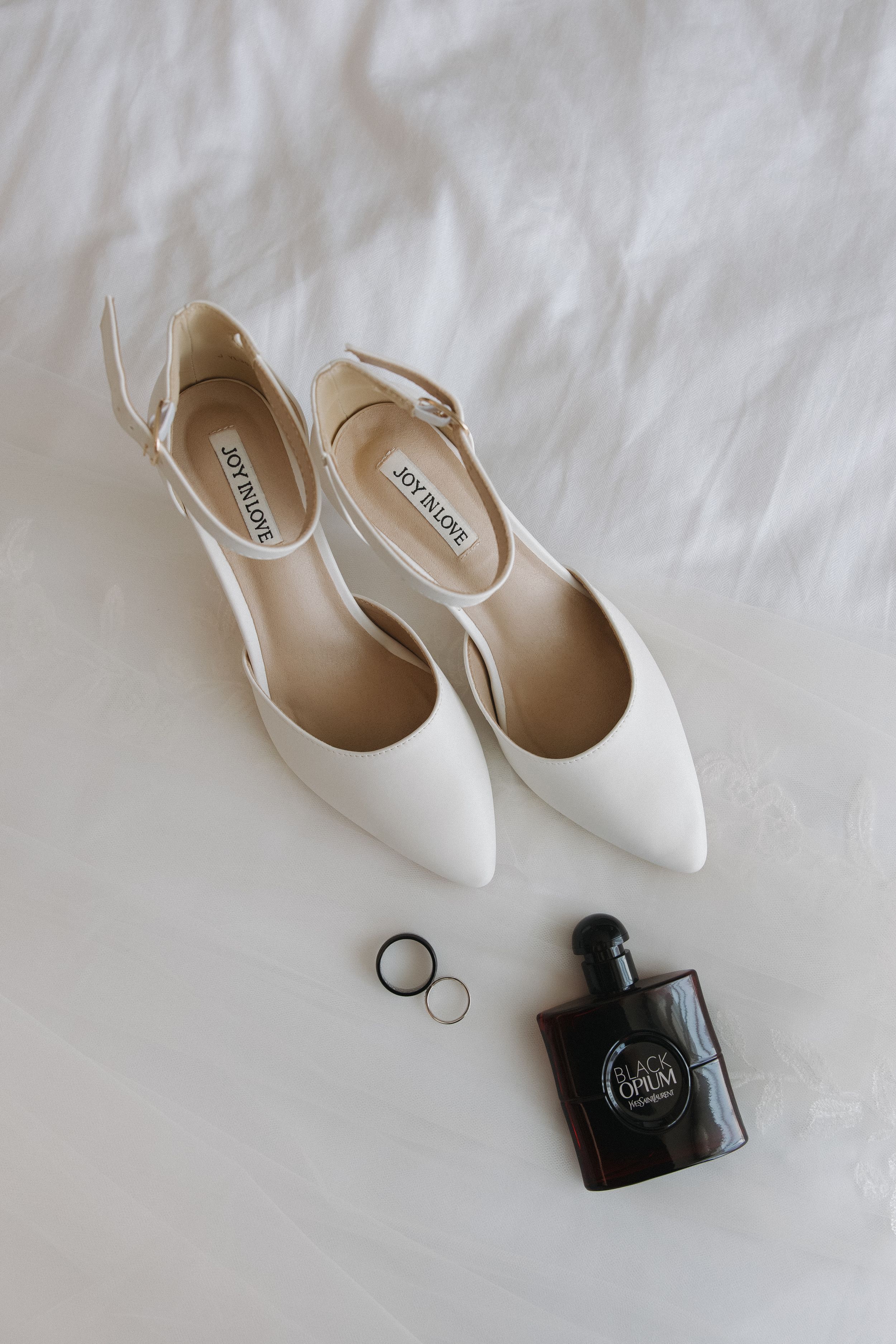 White leather pumps and black wedding rings arranged on a clean white textured surface for a minimalist bridal styling.