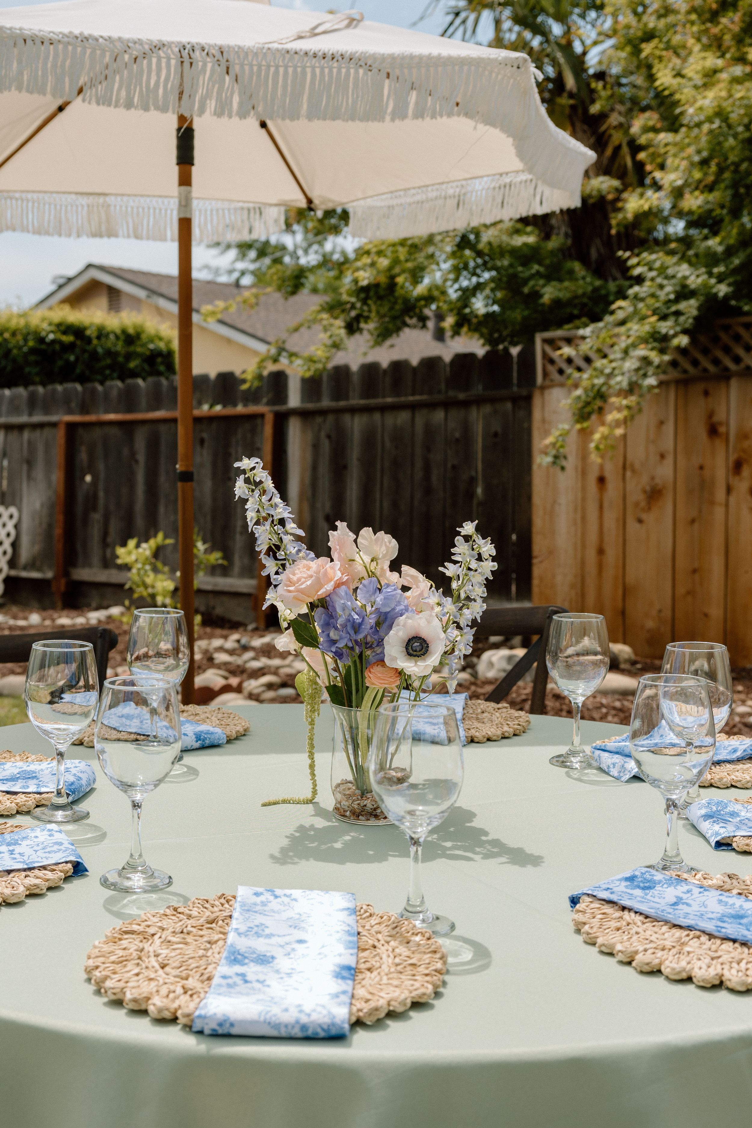 Elegant outdoor table setting with sage green tablecloth, woven placemats and delicate floral centerpiece under white umbrella.