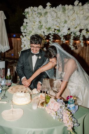 A couple cuts their white wedding cake together at their reception table decorated with pink and white flowers.