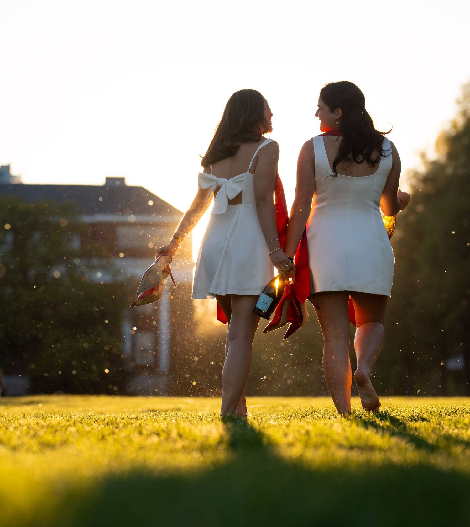 Two women in white dresses walk through a sunlit park, holding red graduation regalia at golden hour.