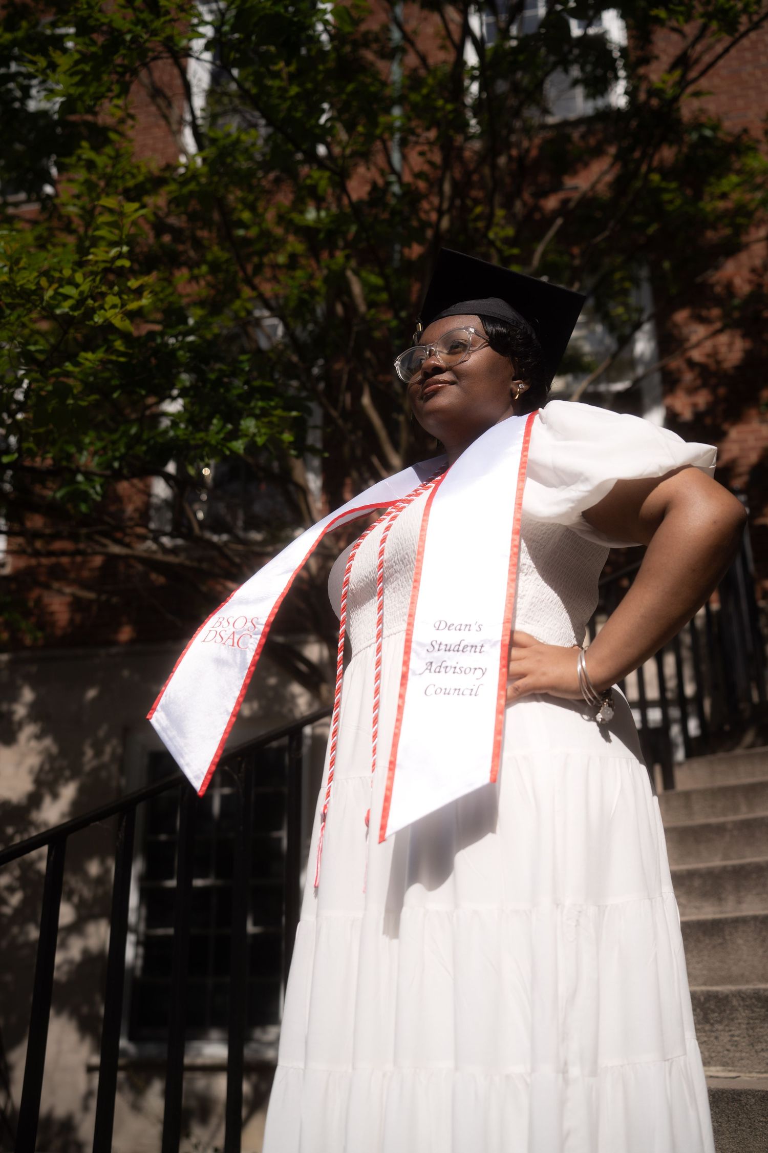 A white graduation gown billows in the wind while being photographed on outdoor steps.