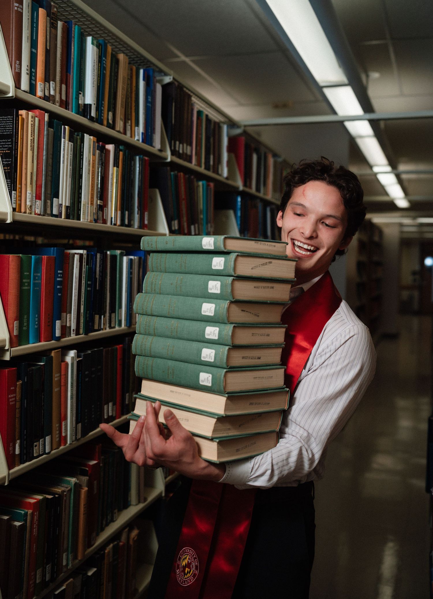 Smiling graduate in red sash holds tall stack of green books in library aisle during graduation photo.