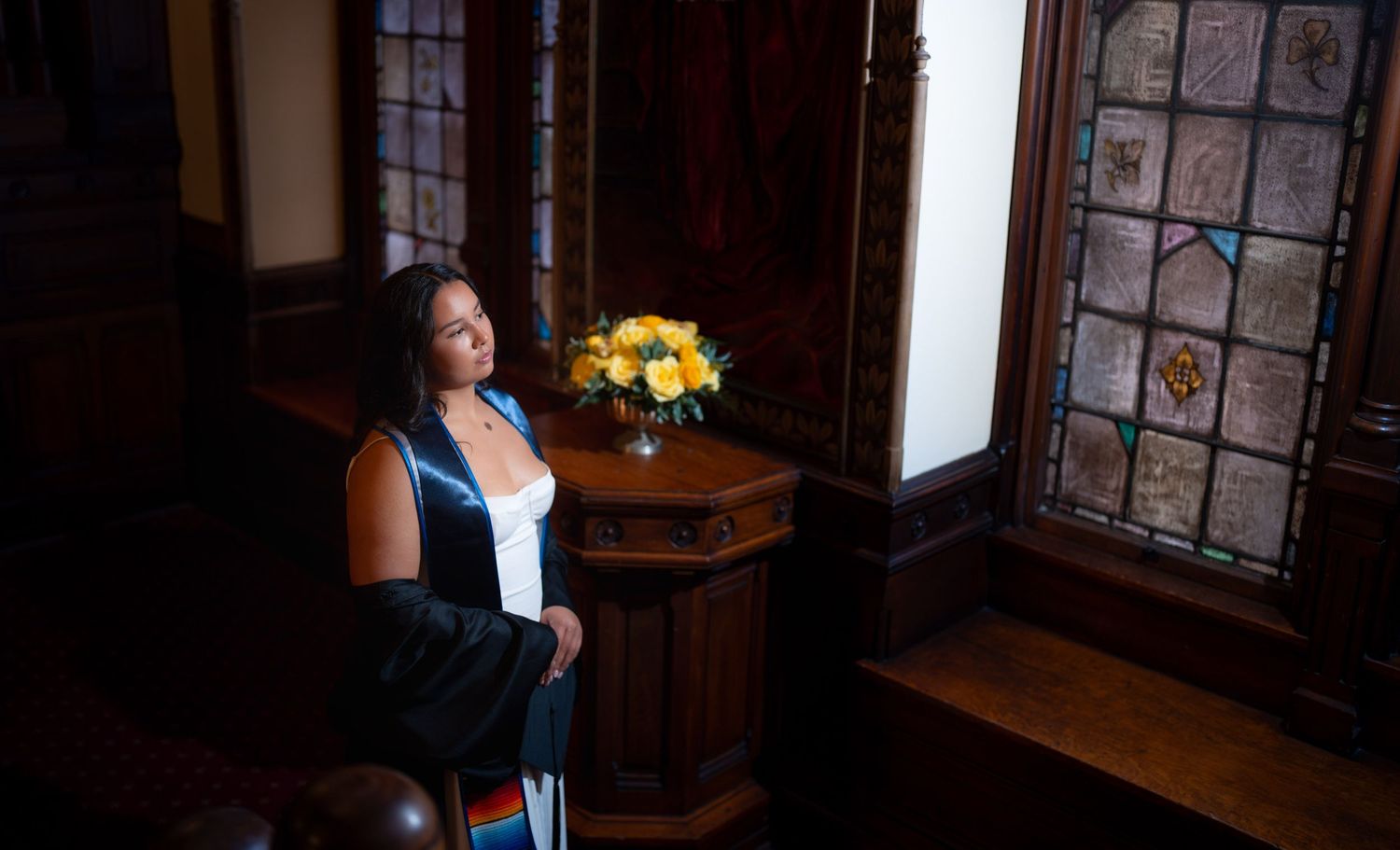 Woman in graduation regalia sits pensively near stained glass window with yellow flower bouquet.