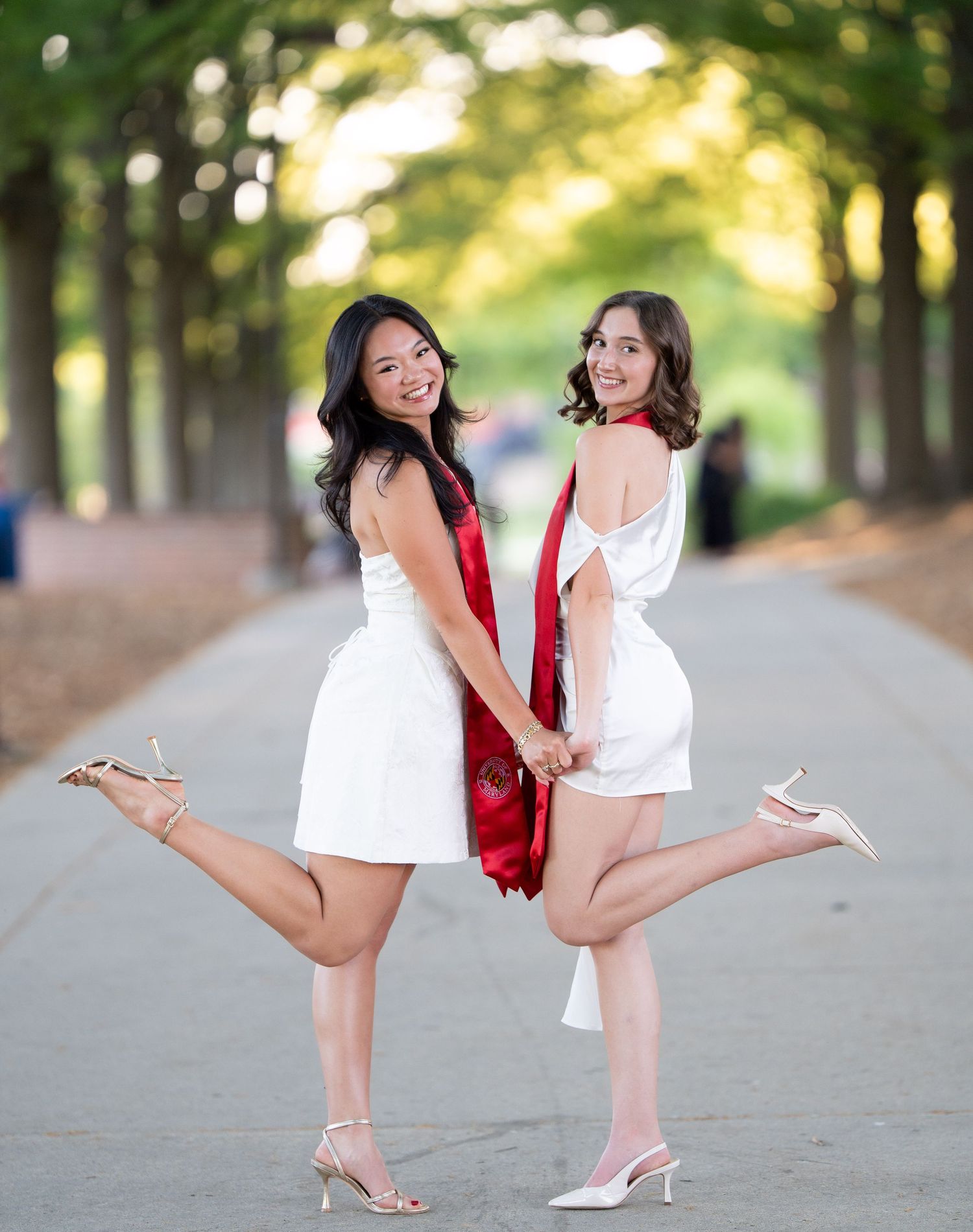 Two smiling graduates in white dresses holding red stoles pose back-to-back on a tree-lined path.