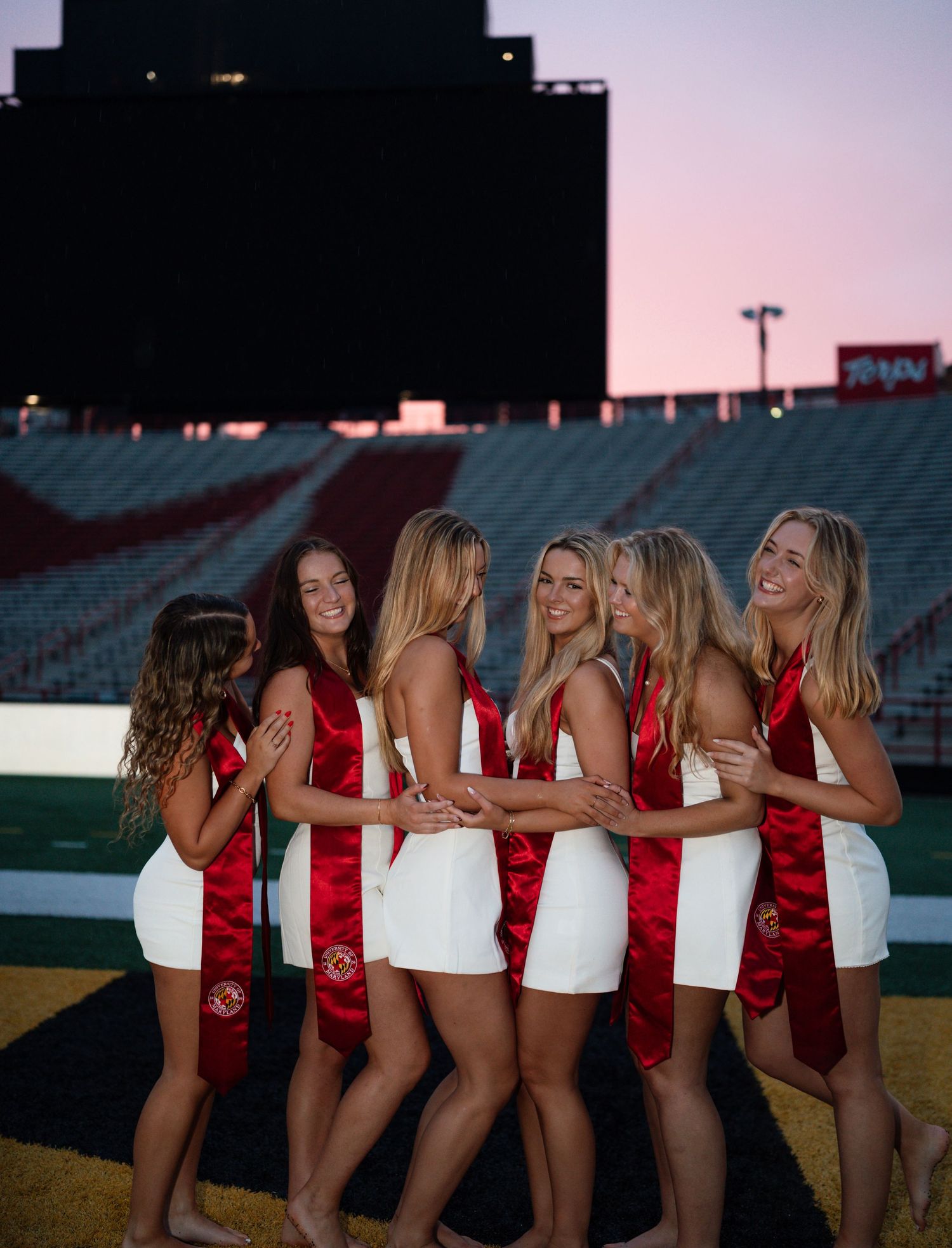 Six young women in white dresses and red graduation sashes pose together on a football field at sunset.