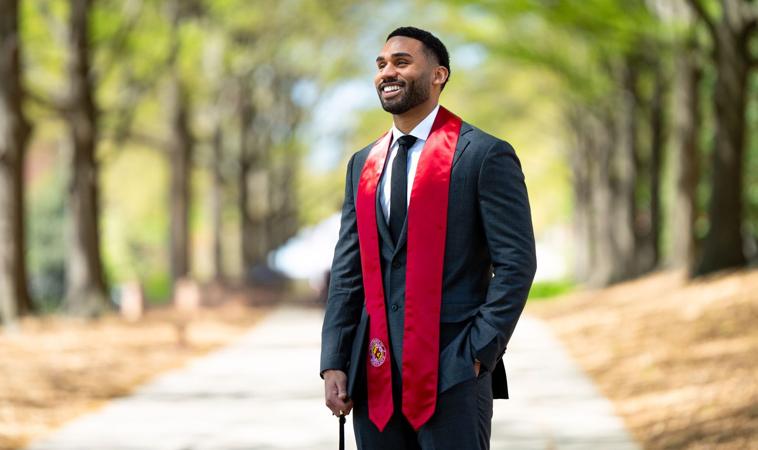 Young man in graduation stole and suit smiling outdoors on a tree-lined path celebrating his achievement.