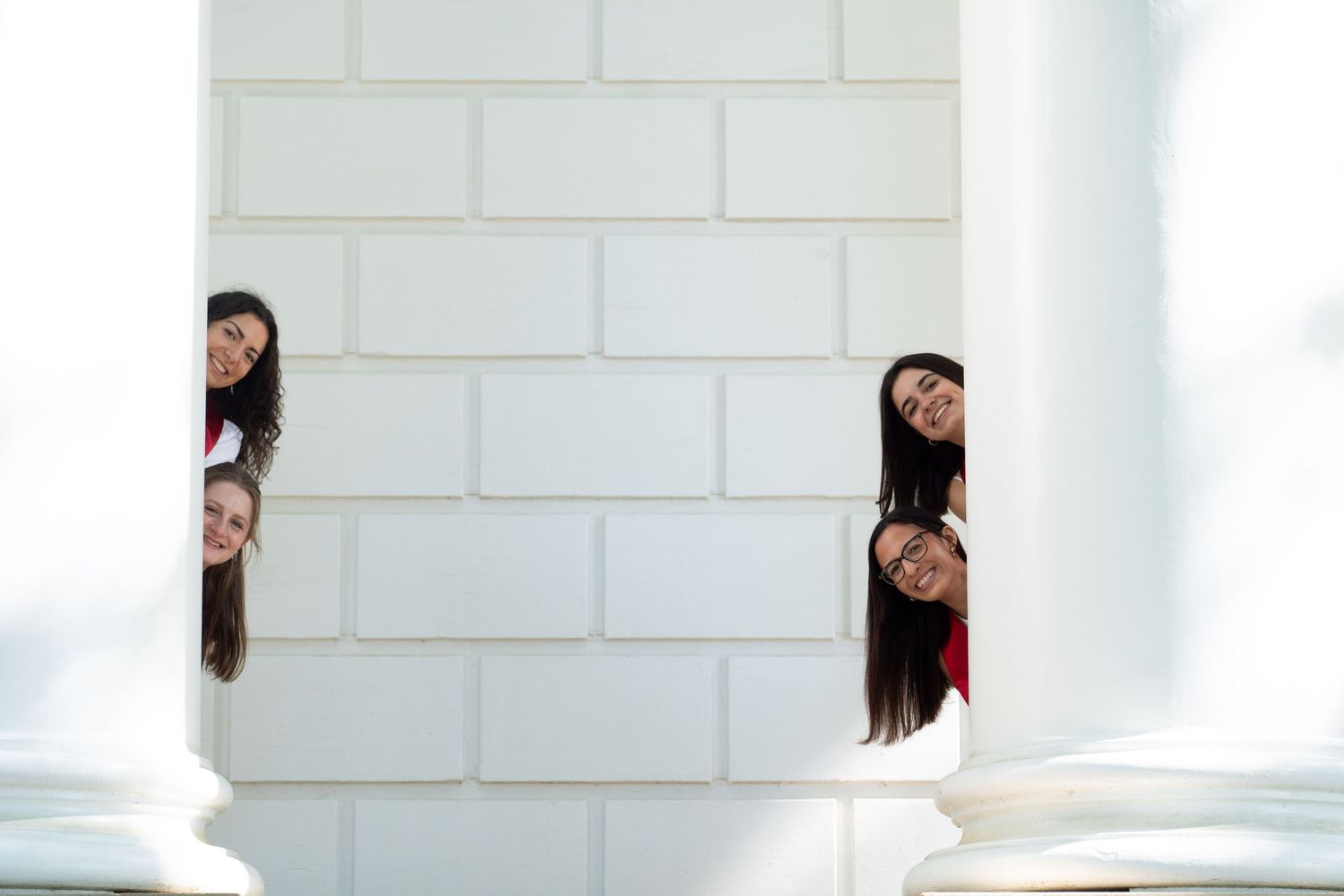 Four young women playfully peeking around a white brick wall column, smiling and posing together outdoors.