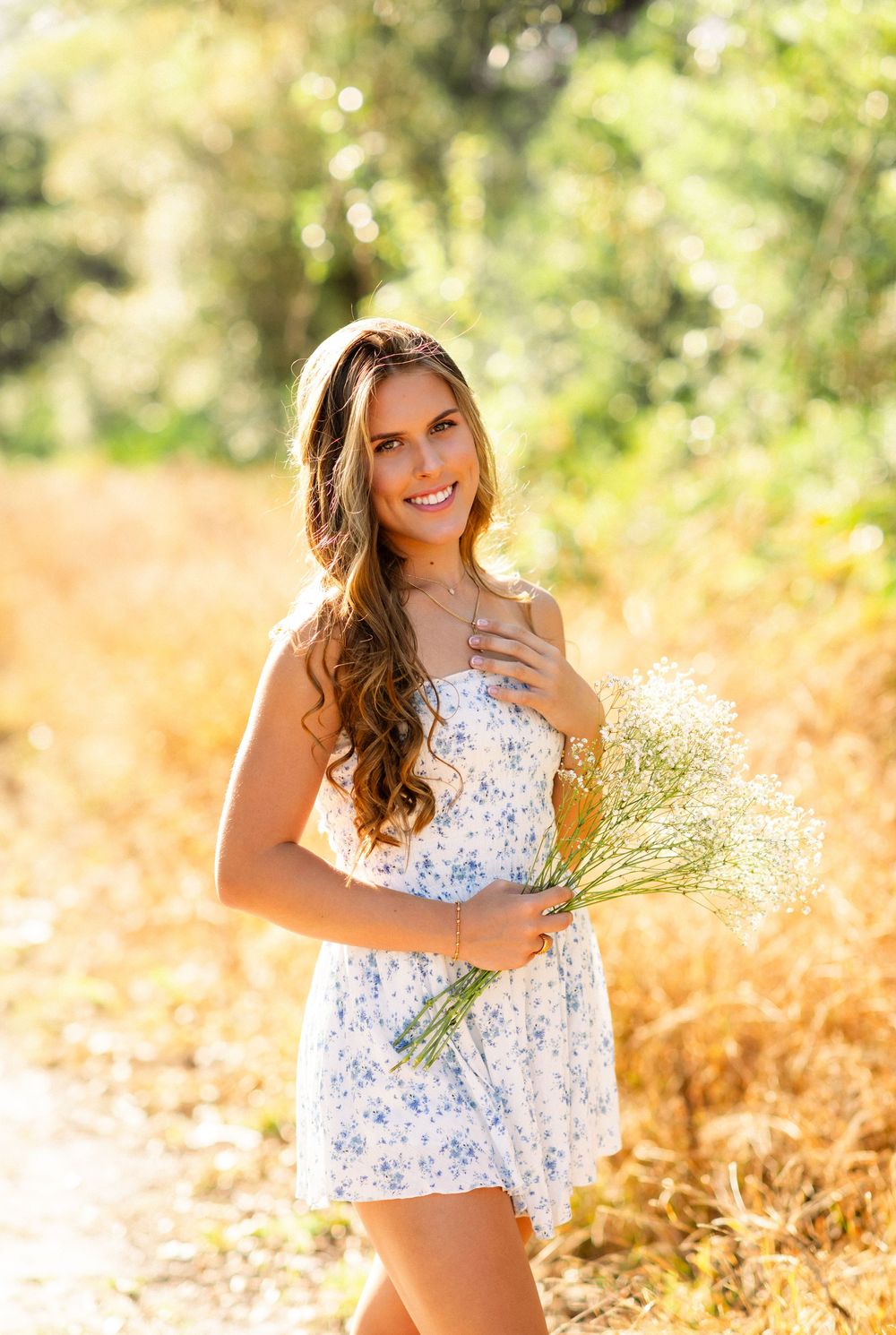 A person in a white floral dress poses gracefully in a sunlit field with golden ferns and warm autumn colors.