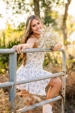 Woman in floral dress poses with baby's breath bouquet by rustic metal fence at golden hour sunset.