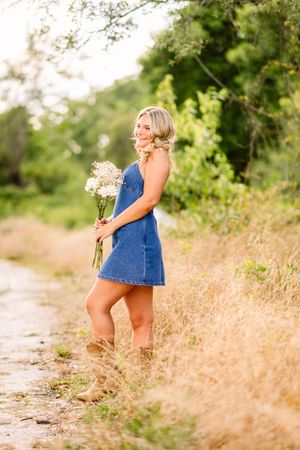 Person in denim dress holding wildflowers poses outdoors in tall golden grass along rural dirt path in summer.