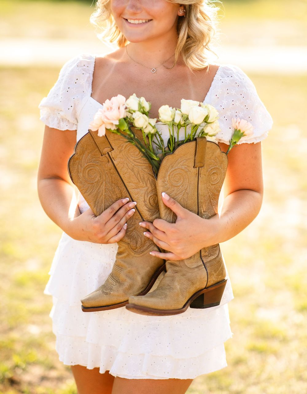 A pair of brown cowboy boots filled with white roses held by someone wearing a white eyelet dress in golden sunlight.