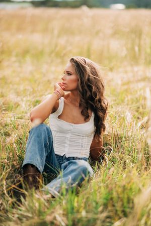 Someone in a white top and jeans sits thoughtfully in a sunlit meadow of tall grass during golden hour.