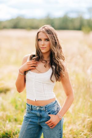 A woman poses in a white crop top and jeans while standing in a sun-lit golden grain field during sunset.