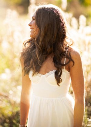 A joyful person in a white flowing dress smiles in golden sunlight while surrounded by tall grass and wildflowers.