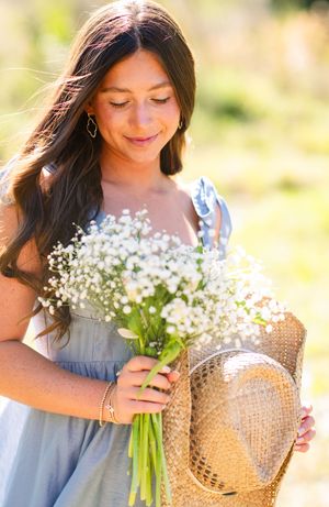 Woman in blue dress holding bouquet of baby's breath flowers and straw hat on sunny summer day.