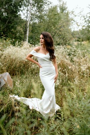 Bride in fitted white dress poses in field of wild summer grasses and flowers.