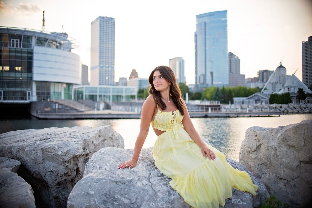 Young woman in a yellow gown sits on rocks with a city skyline at sunset.