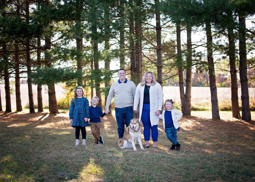 Family of four with a dog posing among tall pine trees on a sunny day.