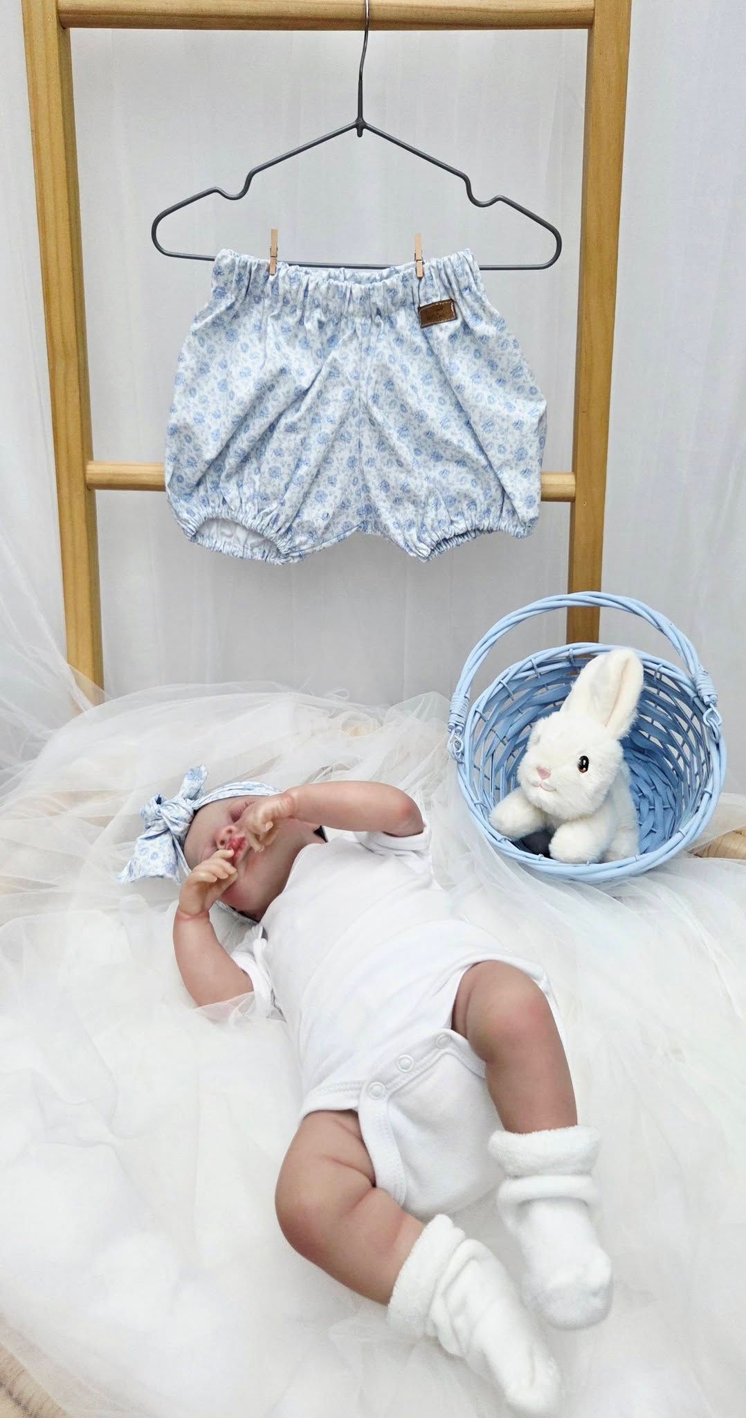 Baby girl lying next to a blue basket with bunny plush, wearing white outfit with floral bloomers displayed on hanger above.